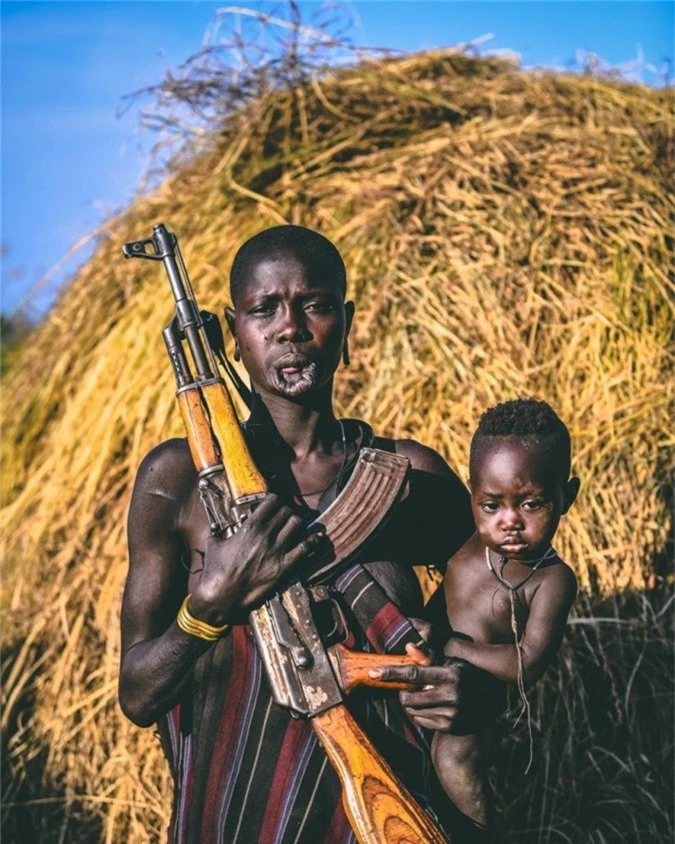 Người phụ nữ bộ lạc Mursi một tay cầm khẩu súng AK-47 và một tay bế con. Súng thường được sử dụng như một vật dụng truyền thống để họ có thể tự bảo vệ mình trước những xung đột bộ tộc.