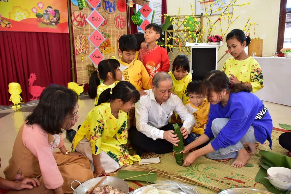 Chủ tịch tỉnh Thừa Thiên Huế ngồi xuống chiếu bắt tay gói bánh chưng, bánh tét cùng cô cháu Làng trẻ em SOS Huế. 